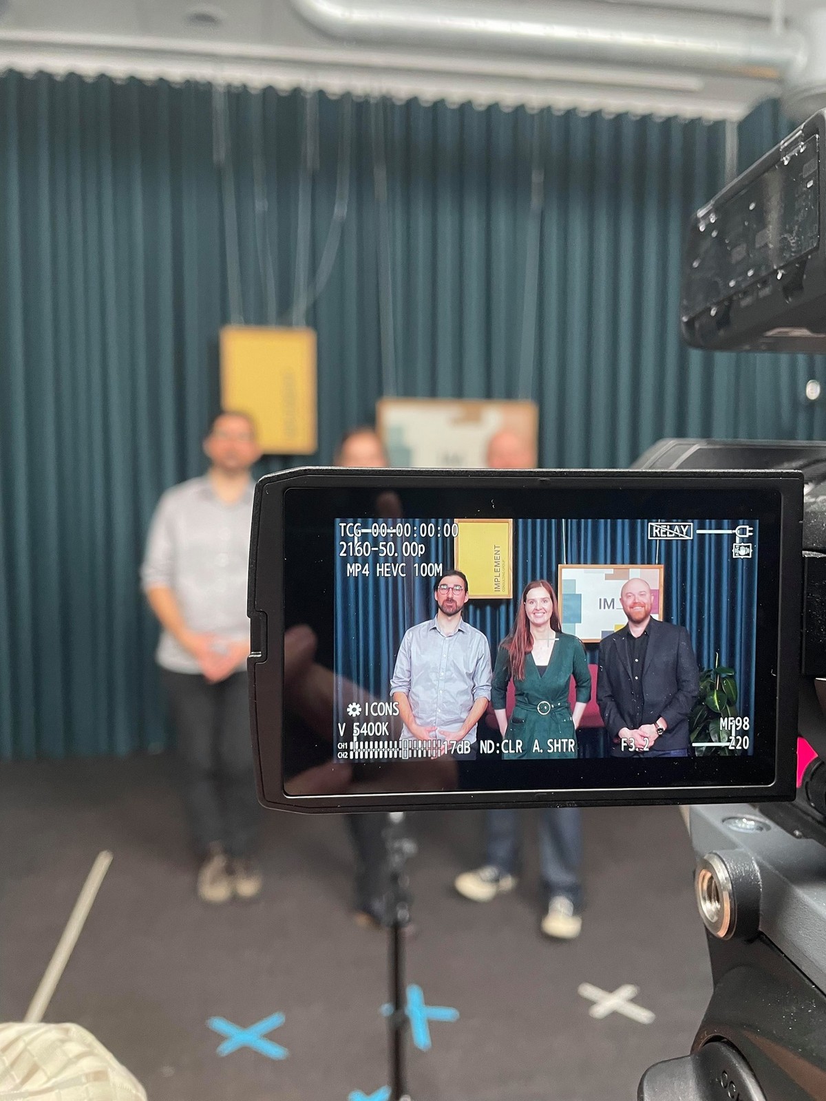 Photograph taken behind a video camera screen that has 3 people standing smiling